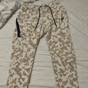 Nike camo mjukisbyxor med snörning - Beiga och vita mjukisbyxor från Nike med camouflagemönster, svarta dragsnören och logga på låret. Byxorna har elastisk midja och praktiska fickor, samt en modern tapered passform. Perfekta för chill eller streetwear.