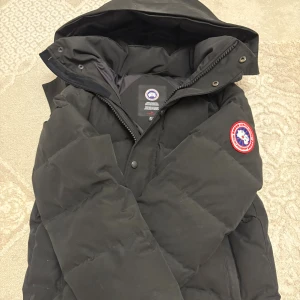 Svart dunjacka från Canada Goose M - Svart dunjacka från Canada Goose med huva och klassisk logga på ärmen. Jackan är quiltad och har dragkedja samt knappar framtill. Perfekt för kalla dagar och har en clean, stilren look som funkar till allt.