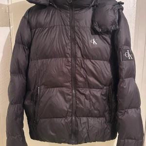 Svart pufferjacka från Calvin Klein Jeans - Svart pufferjacka från Calvin Klein Jeans med huva och dragkedja framtill. Jackan har två fickor med dragkedja och tydliga logodetaljer på bröstet och ärmen. Perfekt för kalla dagar och riktigt snygg streetstil.