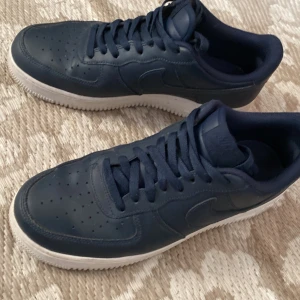 Nike Air Force 1  - Säljer ett par klassiska Nike Air Force 1 i marinblått skinn med vit sula. Skorna har rund tå, snörning och den ikoniska Air Force-sulan. Perfekta för dig som gillar stilrena och tidlösa sneakers med en clean vibe.