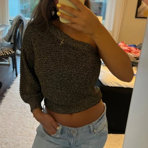 Glittrig oneshoulder stickad topp - Säljer en mörkgrå glittrig stickad topp med oneshoulder-design. Toppen är croppad och har en lång ärm på ena sidan medan andra axeln är bar. As snygg till höst och vinter fester 