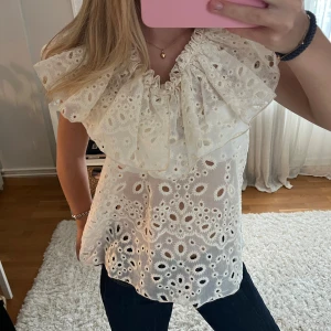 Vit blus med volang och broderi - Supersöt vit blus med stora volanger runt halsen och broderade hålmönster över hela toppen. Blusen är ärmlös och har en luftig, lätt känsla som passar perfekt till jeans eller kjol. 