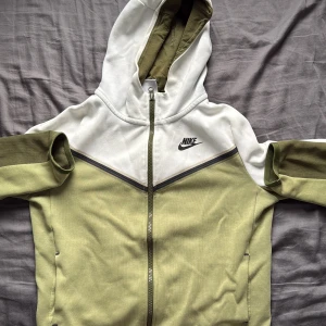 Nike hoodie i vitt och olivgrönt/bluza z kapturem Nike - Nike hoodie med dragkedja i vitt och olivgrönt. Tröjan har huva, långa ärmar och klassisk Nike-logga på bröstet. Snyggt V-format mönster framtill och praktiska fickor. Perfekt för en sportig och avslappnad stil. Den har några skador som du kan se i bilderna 