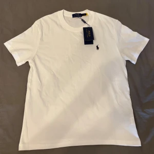 Vit t-shirt från Polo Ralph Lauren - Klassisk vit t-shirt från Polo Ralph Lauren med rund hals och korta ärmar. T-shirten har den ikoniska broderade loggan i svart på bröstet och är gjord i mjuk bomull. Perfekt till jeans eller shorts för en clean och stilren look.