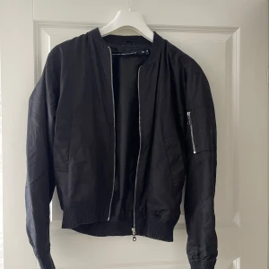 Svart bomberjacka från H&M - Svart bomberjacka med klassisk ribbad krage, muddar och nederkant. Jackan har dragkedja framtill och en extra ficka med dragkedja på ena ärmen. Tillverkad i ett slätt material som ger en clean look. Perfekt för dig som gillar streetstyle.