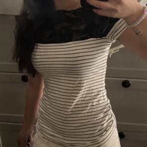 Randig topp med spetsdetalj - Säljer en vit och svart randig topp med tight passform. Toppen har korta ärmar och en snygg svart spets upptill som ger en extra touch. Perfekt för dig som gillar klassiska ränder med en twist.