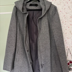 Grå kappa med huva från Vero Moda - Snygg grå kappa från Vero Moda i storlek XL. Jackan har en stor huva, långa ärmar och stängs med både dragkedja och knappar. Materialet är mjukt och känns som ullmix, perfekt för kyliga dagar. Enkel design med diskreta fickor i sidorna. Aldrig använd pga fel storlek🩷