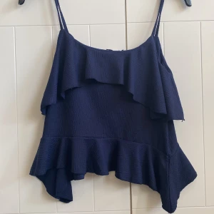 Mörkblå volang linnetopp från Nelly - Säljer en mörkblå linnetopp från Nelly i storlek XS. Toppen har tunna axelband, volangdetaljer både upptill och nedtill samt en lätt ribbad struktur i tyget. Asymmetrisk nederkant ger en cool och unik look. Perfekt för dig som gillar trendiga och feminina plagg.