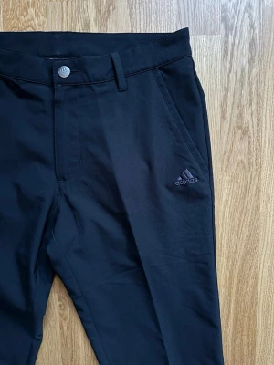 Svarta golfbyxor från Adidas - Snygga golfbrallor från Adidas med logga på fickan, hällor för bälte och fina pressveck. Rak passform, något mindre i storlek (klassiskt adidas) Blandning av bomull och polyester. Dessa är ett måste på golfbanan!