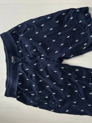 Mörkblå shorts från Polo Ralph Lauren - Mörkblå shorts från Polo Ralph Lauren med vitt broderat logomönster över hela tyget. De har snörning i midjan och är gjorda i mjuk bomull, perfekta för chill dagar. Klassisk och avslappnad passform.