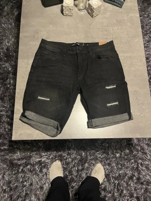 Svarta jeansshorts från Intense - Säljer ett par svarta jeansshorts från Intense i storlek Large. De har slitna detaljer framtill, uppvikta benslut och klassiska fem fickor. Materialet är bomull och de har en normal passform. Perfekt för dig som gillar en lite tuffare stil.