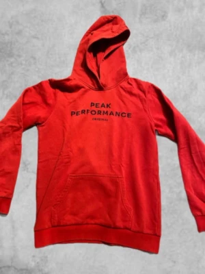 Röd hoodie från Peak Performance - Riktigt snygg röd hoodie från Peak Performance med svart broderad logga på bröstet. Klassisk känguruficka framtill, huva och ribbade muddar. Perfekt för chill dagar eller när du vill ha en sportig look. Storlek 170.