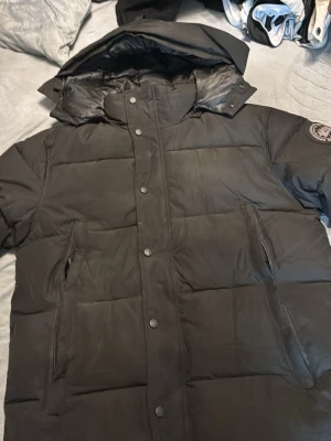 Svart pufferjacka från Canada Goose - Säljer en svart pufferjacka från Canada Goose med huva och emblem på ärmen. Jackan har knappar och dragkedja framtill, två stora fickor och quiltad design. Perfekt för kalla dagar och har en riktigt schysst look.