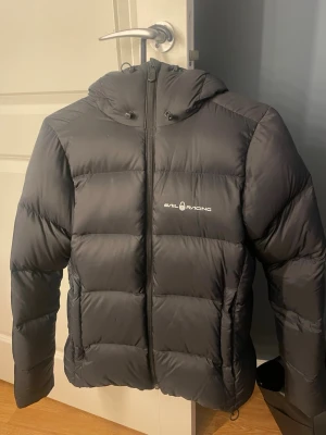 Svart pufferjacka från Sail Racing - Svart pufferjacka från Sail Racing med huva och dragkedja framtill. Jackan har ett stilrent, quiltat mönster och logga på bröstet. Knappt använd och nypris är 4000kr!