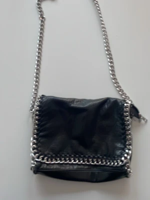 Svart axelväska med kedjedetaljer - Cool svart axelväska i skinn med grova silverfärgade kedjor runt kanterna och som axelrem. Väskan har magnetknäppning och dragkedja på insidan, perfekt för att hålla koll på dina grejer. Stilren design med en edgy touch.