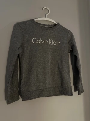 Grå långärmad tröja Calvin Klein - Säljer en stilren grå långärmad tröja från Calvin Klein med logga tryckt framtill. Tröjan har rund halsringning och är tillverkad i mjukt material som passar perfekt till vardags. Enkel och snygg design som funkar till det mesta.