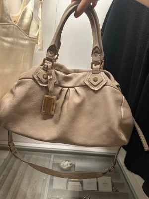 Classic Q groove väska från Marc by Marc Jacobs - Säljer min galet snygga handväska i skinn från Marc by Marc Jacobs med guldiga metalldetaljer och dubbla handtag. Väskan har en avtagbar axelrem och dragkedja upptill. Väskan har en liten fläck på insidan som syns på sista bilden. I övrigt är väskan mycket sparsamt använd!!