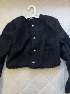 Svart kavajjacka med pärlknappar H&M  - Snygg svart kavajjacka från CICICI med grov textur och croppad passform. Jackan har rund hals, långa ärmar och fyra pärlknappar framtill. Fransen längs kanterna ger en cool vibe och materialet känns lyxigt och lite tjockare.