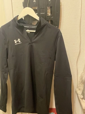 Svart Under Armour träningströja M - Säljer en svart långärmad träningströja från Under Armour i storlek M. Tröjan har en half zip framtill och är gjord i ett mjukt syntetmaterial som andas. Perfekt för sport och aktiva dagar. Logga på bröstet och stilren design.
