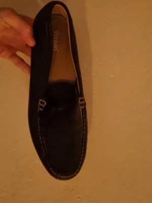 Svarta loafers från Weejuns - Klassiska svarta loafers från Weejuns med dekorativ söm runt tån och diskret rosettdetalj framtill. Skorna har en platt sula i läder och ovandel i skinn, perfekt för en stilren och tidlös look. Passar 43-45