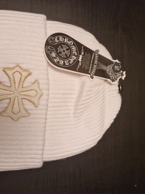 Vit ribbad mössa Chrome Hearts - Säljer en vit ribbad mössa från Chrome Hearts med ett stort broderat kors framtill i vitt och guld. Mössan har en klassisk uppvikt kant och är tillverkad i ett mjukt, stickat material. Perfekt accessoar för dig som gillar streetwear och vill sticka ut.