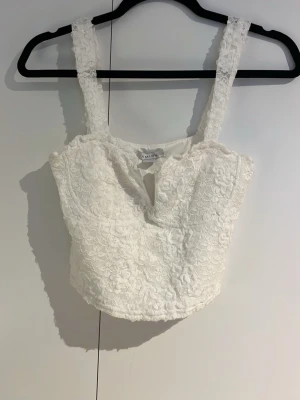 Vit spetstopp - Supersöt vit topp  med breda axelband och blommig spets över hela toppen. Croppad modell med liten v-ringning framtill. Perfekt till sommarens alla tillfällen och ger en romantisk vibe till din outfit.