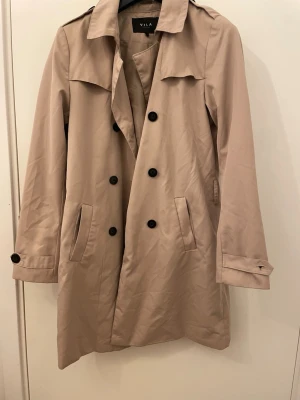 Beige trenchcoat från Vila, stl M - Säljer en klassisk beige trenchcoat från Vila i storlek M. Jackan har dubbelknäppning med svarta knappar, krage och fickor på sidorna. Ärmarna har snygga detaljer med knapp. Perfekt för vår och höst när du vill ha en stilren look. Det fattas en knapp på vänster ärm. Priset går att diskutera. 