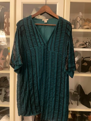 Turkos blus med paljettdetaljer från H&M - Säljer en turkos blus från H&M i storlek M. Blusen har svarta paljettdetaljer och pärlor i ett randigt mönster, v-ringad hals och korta ärmar. Materialet är lätt och har en glansig finish som ger en cool look. Perfekt för dig som gillar att sticka ut.