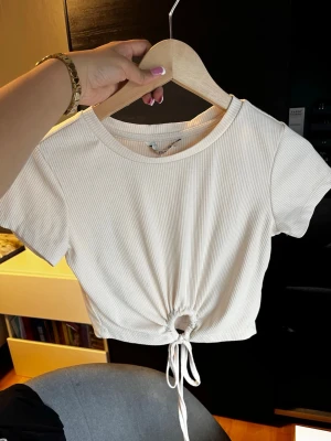 Vit ribbad croppad topp från Bare - Säljer en vit croppad topp från Bare med korta ärmar och rund hals. Toppen är ribbad och har en snygg snörning framtill som ger en cool detalj. Perfekt för sommarens alla häng! Materialet känns mjukt och stretchigt.