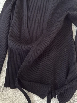 Svart omlottopp från Soaked in Luxury - Säljer en svart ribbad omlottopp från Soaked in Luxury i storlek S. Toppen har långa ärmar, v-ringad hals och knytband i midjan. Perfekt för dig som gillar stilrena och bekväma plagg med snygg passform.