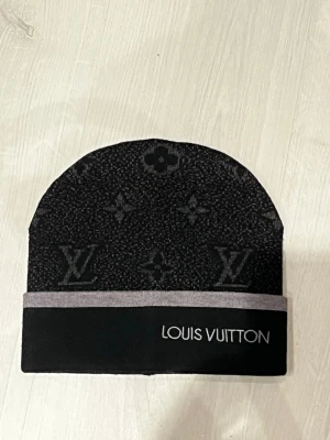 Svart Louis Vuitton mössa - Säljer en svart mössa från Louis Vuitton med klassiskt monogrammönster och grå kant. Mössan har en stilren design med LV-logga och är perfekt för dig som vill ha något exklusivt och snyggt till vintern. Använd endast en gång . Fraktar samma dag!