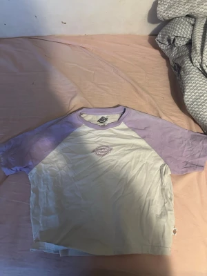 Ljuslila och vit Dickies t-shirt - Säljer en snygg t-shirt från Dickies med ljuslila ärmar och vit kropp. Klassisk raglanmodell med tryckt Dickies-logga på bröstet. Perfekt för dig som gillar streetwear och vill ha en chill vibe. Gjord i mjuk bomull för skön känsla hela dagen.