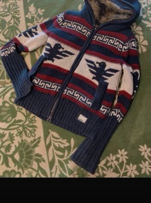 Blå stickad hoodie från Superdry - Säljer en blå stickad hoodie från Superdry med pälsfoder i huvan. Tröjan har mönster i vitt och rött, dragkedja framtill och långa ärmar. Grymma detaljer med fågelmotiv och grafiska ränder. Perfekt för kalla dagar när du vill vara både varm och snygg.