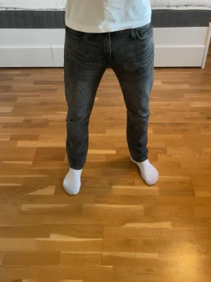 Jack&Jones jeans med slitningar  - Säljer ett par mörkgrå jeans från Jack & Jones med slitningar och hål på ena benet. Modellen har klassisk femficksdesign, normal passform och raka ben. Jeansen är i ett mjukt denimtyg och har en snygg tvättad look.