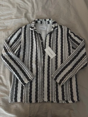 Randig skjorta från Ciszere - Säljer en snygg skjorta från Ciszere i vit och svart med vertikala ränder. Skjortan har krage, knappar framtill och lång ärm. Materialet är stickat och känns riktigt chill. Perfekt för dig som gillar unika detaljer och mönster.