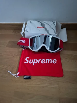 Supreme x Smith skidglasögon - Supreme x Smith skidglasögon med vit ram och spegelglas. Rött band med Supreme-logga i vitt och justerbar passform. Kommer med Supreme-fodral och originalkartong med marmormönster. Perfekt för dig som vill sticka ut i backen.                                          Vid minsta fråga är det bara skicka ett meddelande!