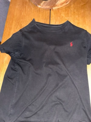 Svart t-shirt från Ralph Lauren - Svart t-shirt från Ralph Lauren med klassisk röd logga broderad på bröstet. Modellen är kortärmad och har en rund halsringning. Tillverkad i mjuk bomull som känns skön mot huden. Perfekt för dig som gillar stilrena och enkla plagg med en touch av lyx.