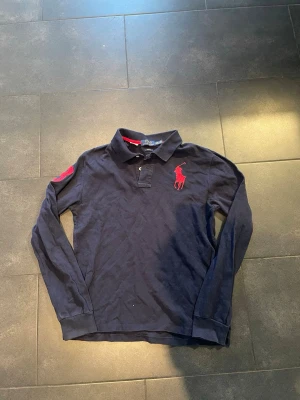 Mörkblå långärmad piké från Polo Ralph Lauren - Snygg mörkblå långärmad pikétröja från Polo Ralph Lauren med röd broderad logga på bröstet och en stor röd siffra 3 på ärmen. Slim fit-modell med klassisk krage och knappar framtill. Perfekt för dig som gillar sportig och stilren look.