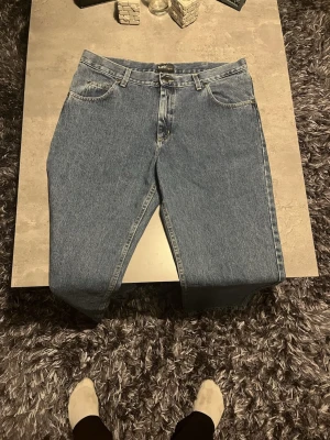 Blå jeans från Lee med raka ben - Säljer ett par klassiska blå jeans från Lee med raka ben och normal passform. Jeansen har fem fickor, bälteshällor och Lee-logga på bakfickan. Materialet är robust denim i bomull och färgen är en snygg mellanblå ton. Perfekt för dig som gillar tidlös stil.