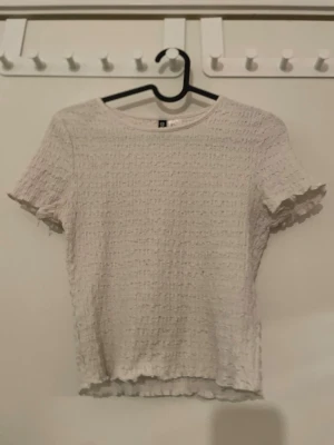 Vit ribbad topp från Divided - Säljer en söt vit topp från Divided i storlek S. Toppen har korta ärmar, ribbad struktur och lätt vågiga kanter vid ärmslut och nederkant. Materialet är mjukt och stretchigt, perfekt till jeans eller kjol.