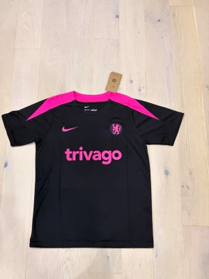 Svarta och rosa Nike Chelsea fotbollströja - Chelsea FC x Nike Dri-Fit fotbollströja i svart med starka rosa detaljer på axlar och krage. Trivago-logga och Chelsea-lejonet i rosa framtill. Tillverkad i lätt polyester som andas, perfekt för träning eller match.