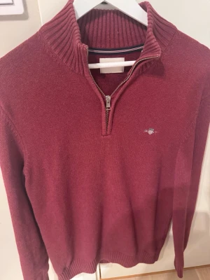 Gant half zip - Säljer en vinröd stickad tröja från Gant med ribbad krage och half zip framtill. Tröjan har ett litet broderat logomärke på bröstet och långa ärmar. Perfekt för dig som gillar klassisk och stilren design.