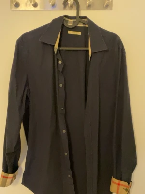 Svart Burberry Brit skjorta M - Svart skjorta från Burberry Brit i storlek M. Klassisk modell med långa ärmar och knappar framtill. Snygga rutiga detaljer i beige och rött på insidan av manschetterna och kragen. Perfekt för dig som gillar stilrena plagg med en twist. Köpt för 1350kr av en vän. Några funderingar på äktheten är det bara att skriva. Pris går diskuteras bara på snabbköp!