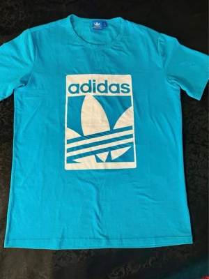 Turkos Adidas t-shirt med stor logga M, 100% polyester  - Snygg turkos t-shirt från Adidas med stor vit logga och text framtill. Klassisk rund hals och korta ärmar. Tillverkad i mjuk bomull som känns skön mot huden. Perfekt för dig som gillar sportig och clean stil.