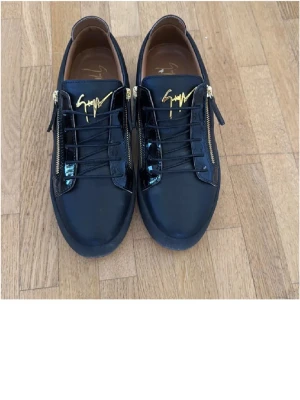 Svarta sneakers från Giuseppe Zanotti - Säljer ett par svarta sneakers från Giuseppe Zanotti med glänsande lackdetaljer, guldfärgade dragkedjor och logga på plösen. Skorna har snörning och en rund tå, tillverkade i skinn och syntet. Riktigt snygga och sticker ut med sina exklusiva detaljer.