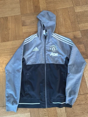 Manchester United vindjacka Adidas - Snygg Manchester United vindjacka från Adidas i grått och svart med huva och dragkedja. Jackan har vita ränder på axlarna, klubbmärke och AON-logga på bröstet. Tillverkad i lätt polyester med meshfoder, perfekt för blåsiga dagar.