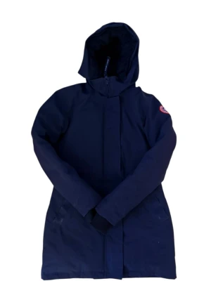 Mörkblå dunparkas från Canada Goose women - Säljer en mörkblå dunparkas från Canada Goose med huva och klassisk logga på ärmen. Jackan har dragkedja och täckande vindslå, långa ärmar och quiltad insida. Perfekt för kalla dagar och har en clean, stilren look.