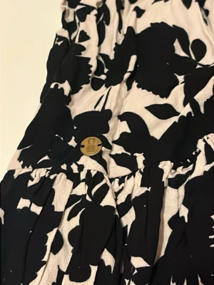 Svartvit blommig kortkjol från Pernilla Wahlgren  - Supersöt kortkjol köpt på Ullared men från Pernilla Wahlgren! Svartvitt blommigt mönster. Kjolen har smock i midjan och volangkant nertill. Tunt, flowy material som känns lätt och luftigt. Perfekt för sommardagar! Använd ett fåtalgånger!