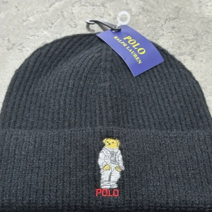 Svart ribbad mössa från Polo Ralph Lauren - Svart ribbstickad mössa från Polo Ralph Lauren med broderad nalle i astronautdräkt framtill och röd POLO-text. Klassisk design med uppvikt kant och mjukt material som håller dig varm. Perfekt accessoar för kyliga dagar.