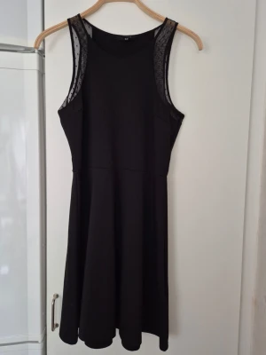Klänning - Svart klänning från H&M i storlek M. Klänningen har ärmlös design med meshdetaljer vid axlarna och en rund halsringning. Kjolen är lätt utsvängd och tyget känns mjukt och följsamt. Perfekt för dig som gillar stilrena och enkla plagg.